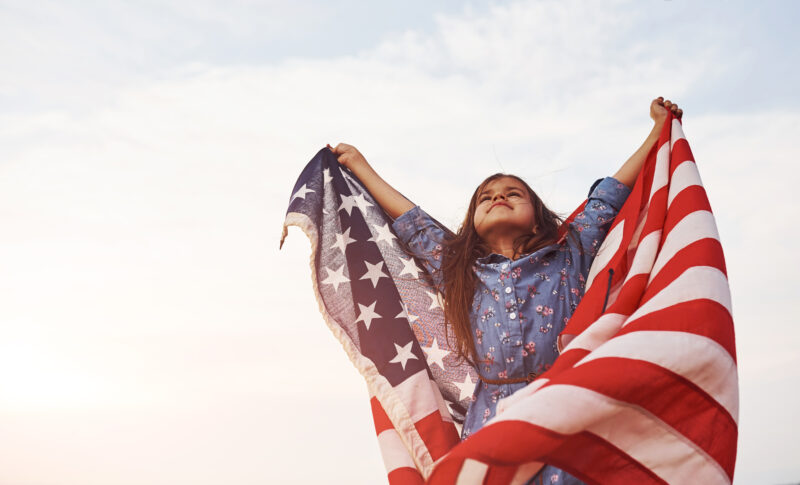 Patriotic female kid with American Flag in hands Against cloudy sky